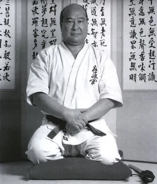 Masutatsu Oyama in Seiza-Haltung mit schwarzem Gürtel, Kyokushin-Gi und Katana vor japanischen Kalligraphie-Schriftrollen