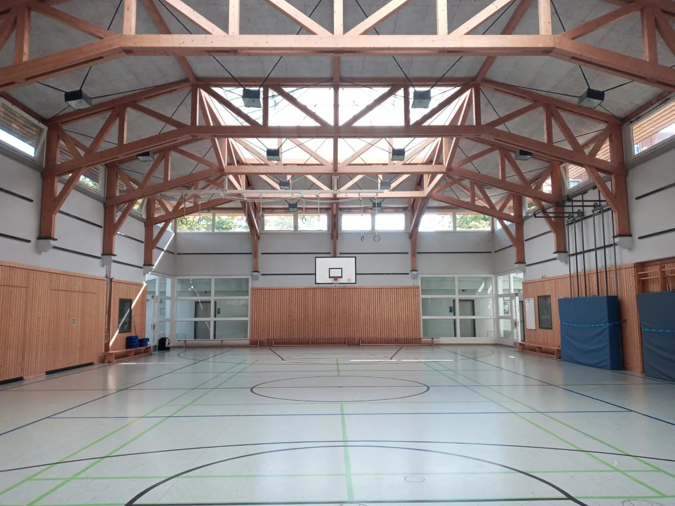 Symmetrische Gesamtansicht der leeren Sporthalle der Edison-Grundschule mit markiertem Hallenboden und markanter Dachkonstruktion aus Holz.