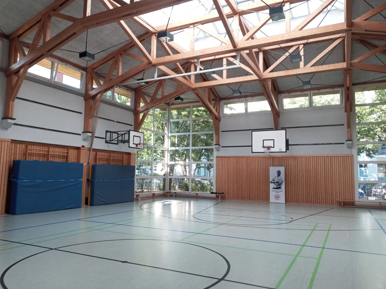 Innenansicht der hellen Sporthalle der Edison-Grundschule mit Holzdeckenkonstruktion, Basketballkörben und einem Karate-Roll-up-Banner des Kyokushinkai Karate Köpenick e.V.