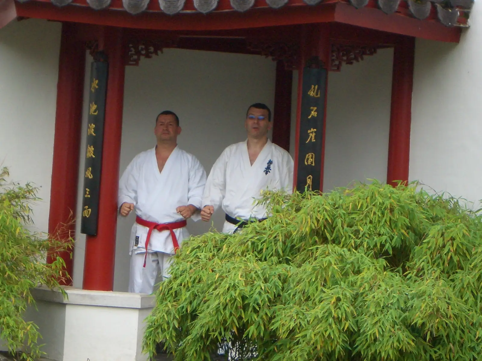 Senpai Peter Bradler und Karateka am chinesischen Pavillon Peter Bradler (Vorsitzender KKK e.V.) mit rotem Gürtel und ein weiterer Karateka mit schwarzem Gürtel stehen vor einem traditionellen chinesischen Schrein-Pavillon