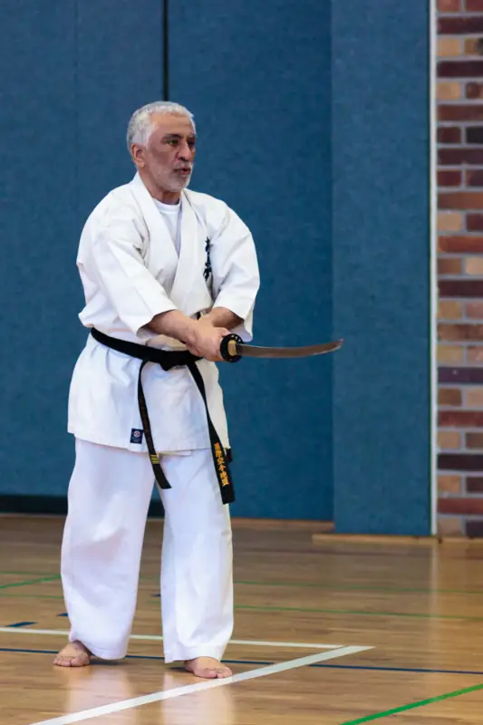 Sensei Moss Vorführung Katana Techniken Lindow 2013 Das Foto zeigt eine Auswahl an traditionellen Kobudo-Waffen des Kyokushinkai Karate Köpenick e.V. (KKK e.V.) an einer Wand in der Sporthalle in Lindow 2013. Zu sehen sind hölzerne Bo-Stäbe, echte Katana in ihren Scheiden, Tonfas sowie Sporttaschen und ein Feuerlöscher im Hintergrund.