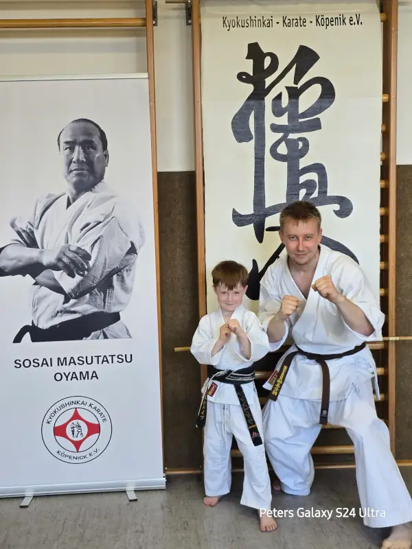 Tim mit Nachwuchs-Karateka – Dojo Briesen Tim und ein Nachwuchs-Karateka des Kyokushinkai Karate Köpenick e.V. in Kampfstellung vor Bannern von Sosai Masutatsu Oyama und dem Vereinslogo
